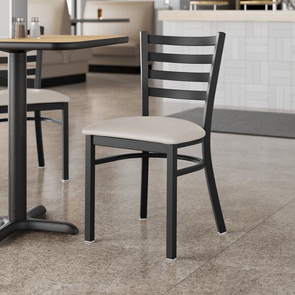 A black Lancaster Table & Seating ladder back chair with a light gray vinyl padded seat at a table in a restaurant.