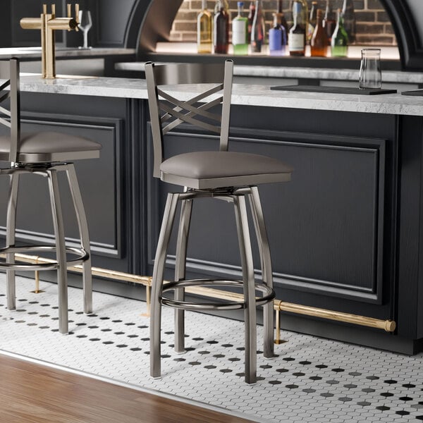 A metal bar stool with a cross back design and a dark gray vinyl padded seat.