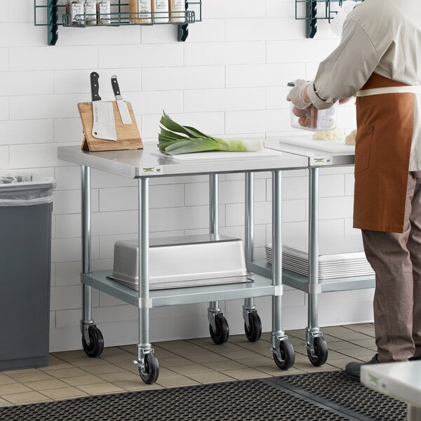 A man in an apron standing next to a Regency stainless steel work table with wheels and an undershelf.