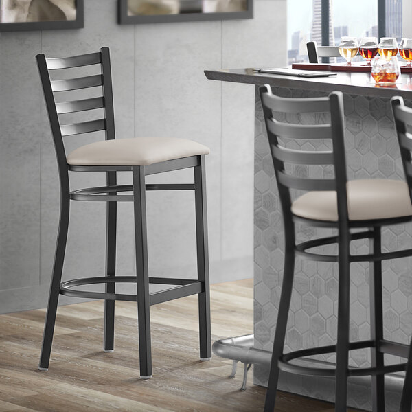 A Lancaster Table & Seating black ladder back bar stool with a light gray padded seat next to a table.