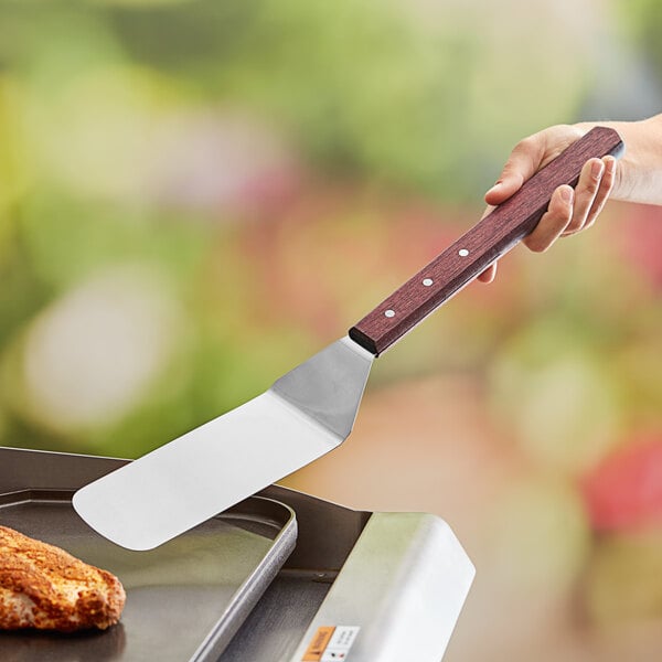 A person holding a Thunder Group solid turner with a wooden handle over a grill.