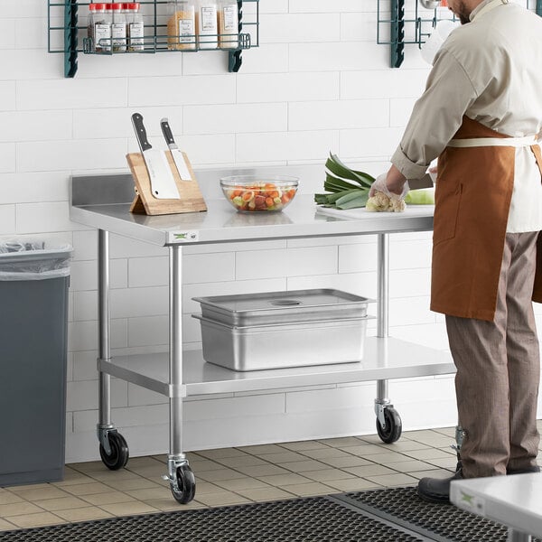 A man in a brown apron cutting vegetables on a stainless steel work table with casters.