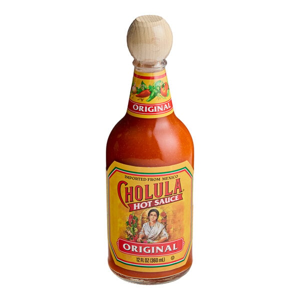 A bottle of orange-red hot sauce with a wooden cap and a yellow label featuring the brand name and an illustration of a woman.