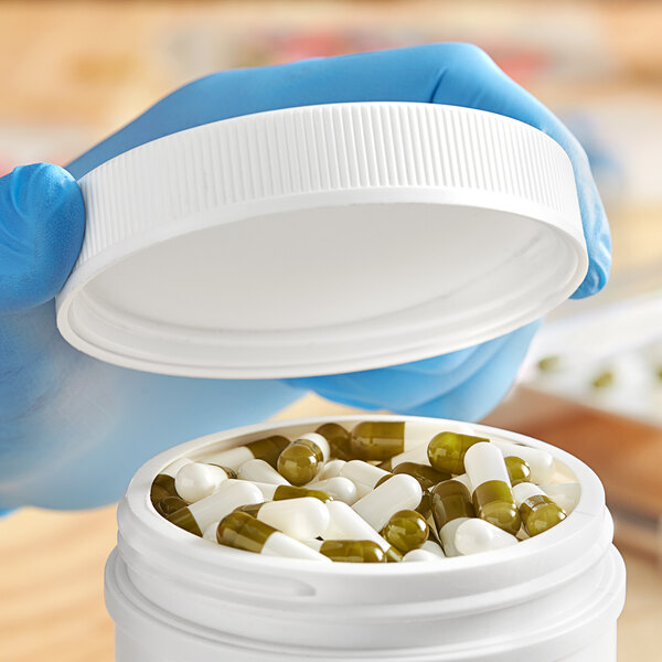 A hand in blue gloves holding a white plastic container with pills.