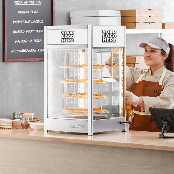 A woman wearing a white apron standing behind a ServIt pizza display case.