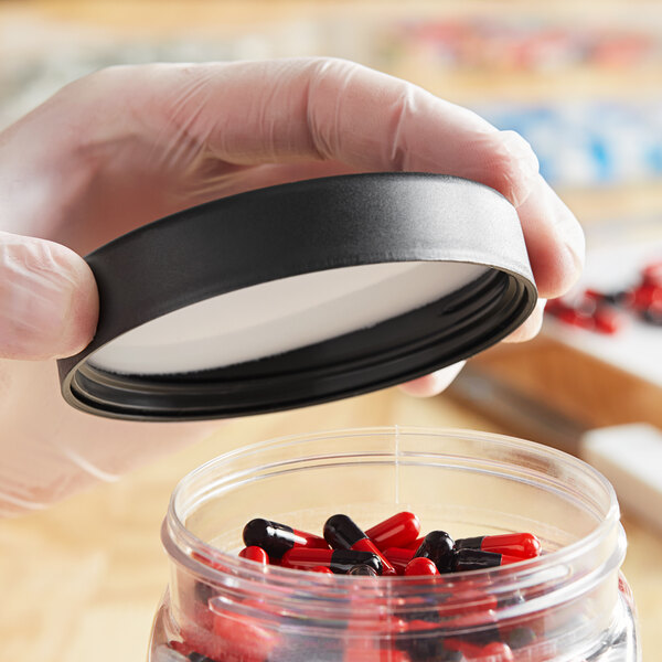 A gloved hand holding a black 89/400 Child-Resistant Pictorial Cap over a container of pills.