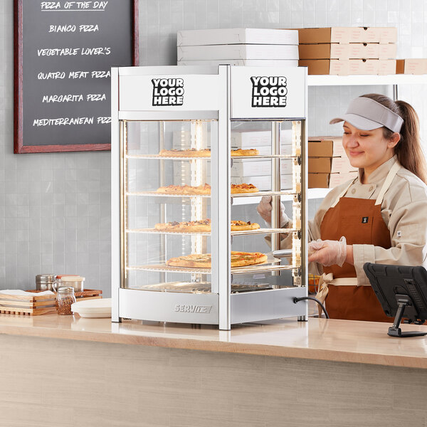 A woman in an apron standing behind a ServIt countertop display case with food.