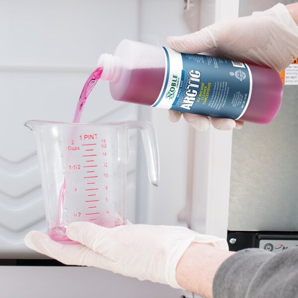 A hand in a white glove pouring pink Arctic Concentrated Ice Machine Cleaner into a measuring cup on a counter.