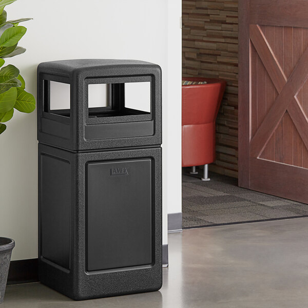 A black Lavex waste container next to a plant.