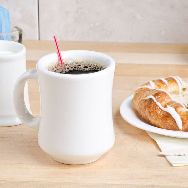 A white Tuxton Princess China mug filled with coffee and a straw on a table with a pastry.