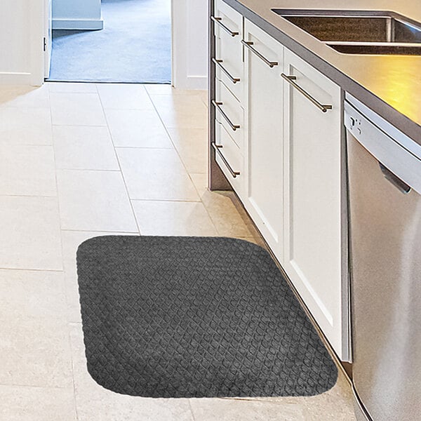 A rectangular, dark gray anti-fatigue mat with rounded corners is placed on a light-colored kitchen floor in front of a sink and cabinets.