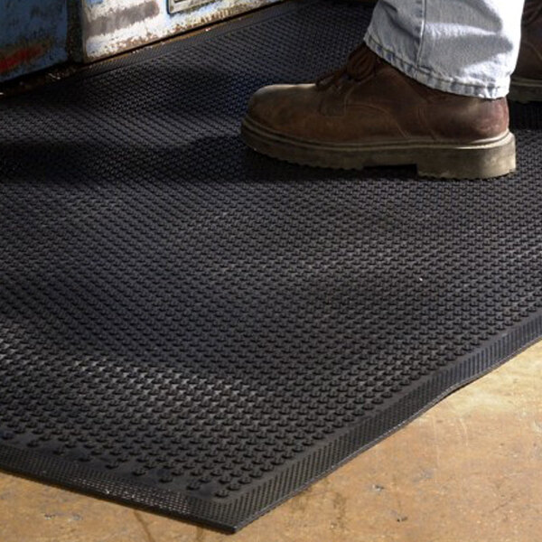 A person wearing brown work boots and light blue jeans is standing on a textured black mat with a slip-resistant surface.