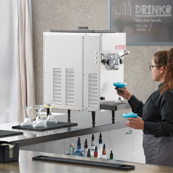 A woman using a SaniServ frozen cocktail machine to fill a glass with blue liquid.