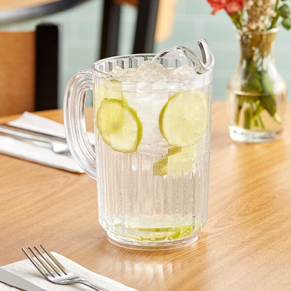 A Carlisle clear polycarbonate beverage pitcher of water with lime slices and ice on a table.