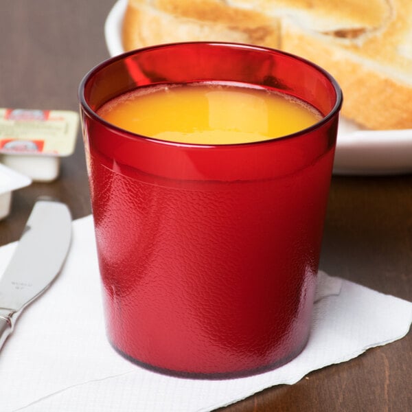 A close-up of a Cambro ruby red plastic tumbler filled with orange juice on a table with bread.