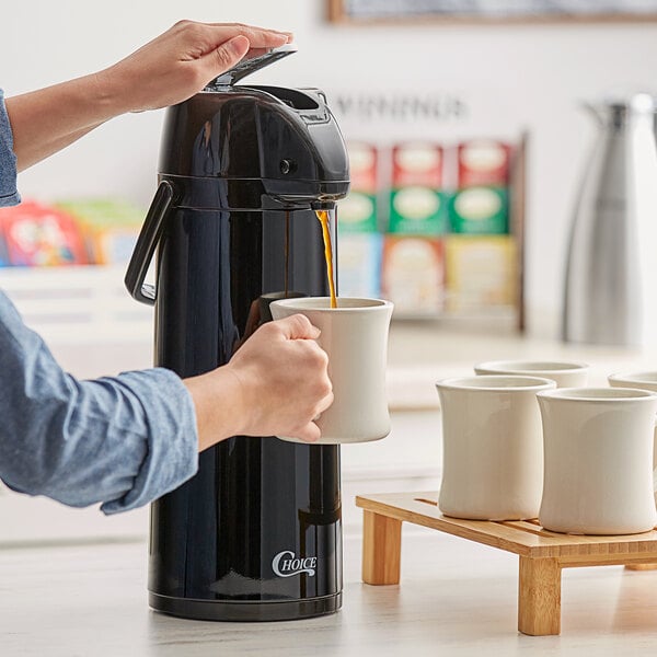A person using a Choice black stainless steel airpot to pour coffee into a white mug.