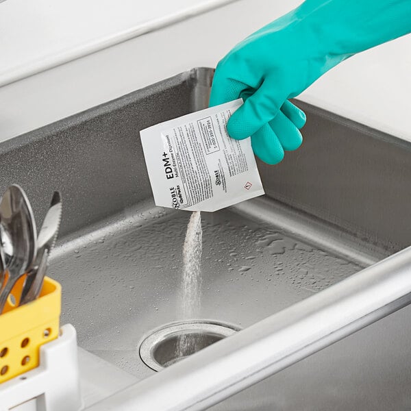 A gloved hand pouring a Noble Chemical EDM+ QuikPack packet into a sink.