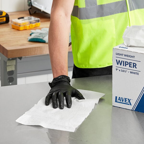 A person wearing a safety vest and gloves cleaning a table with a white Lavex industrial wiper.