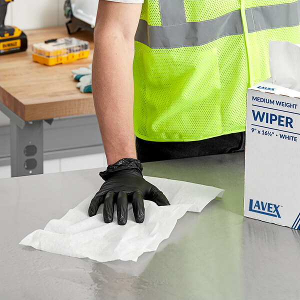 A person wearing black gloves and a safety vest cleaning a table with a Lavex white industrial wiper.