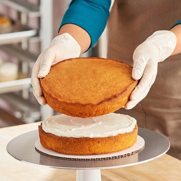 A person assembling a round white layer cake by placing one cake layer on top of another with white icing in between.