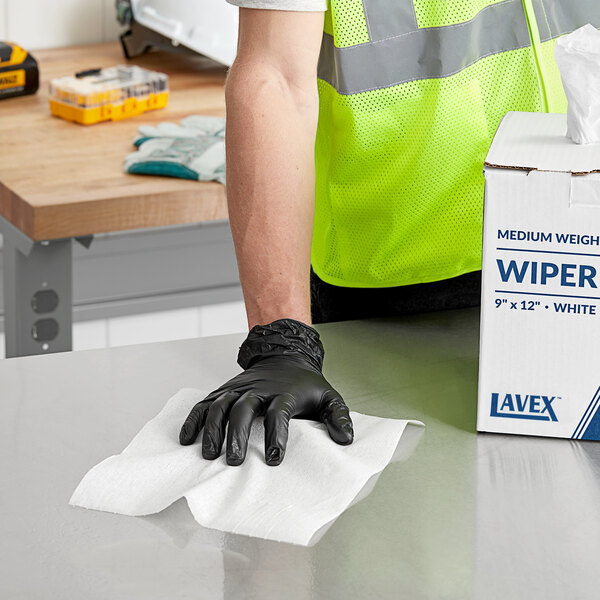 A person wearing a safety vest and gloves pulling a white Lavex industrial wiper from a box.