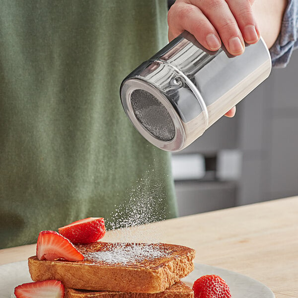 A hand using a Choice stainless steel shaker to sprinkle powdered sugar on French toast.