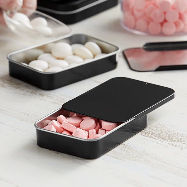 A black tin with slide top filled with pink candies on a table.