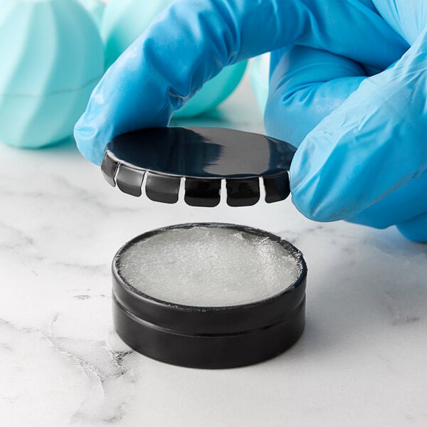 Gloved hands holding a black round container with a black lid containing white substance.