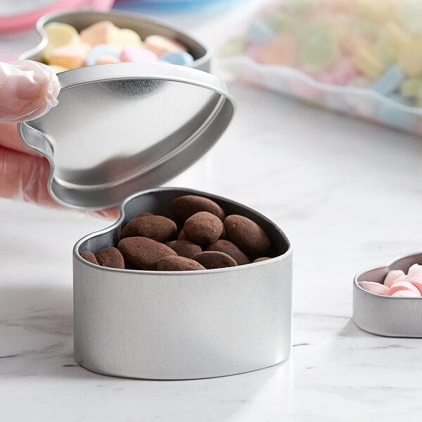 A hand holding a silver heart-shaped tin filled with chocolate candies.