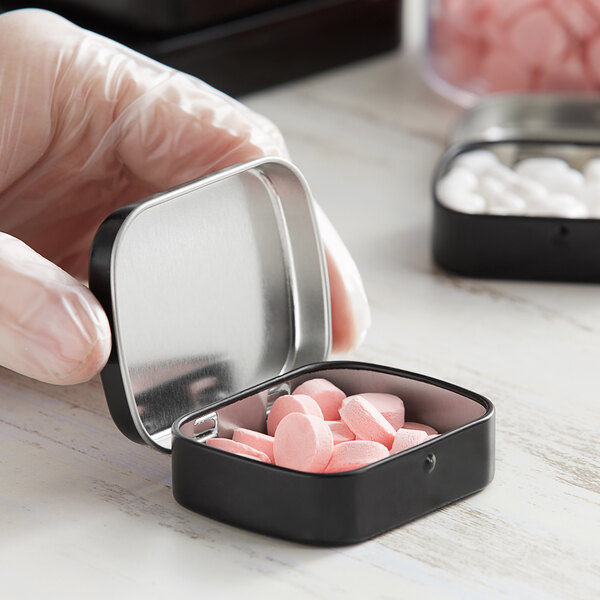 A hand holding a small black metal rectangular tin filled with pink pills.