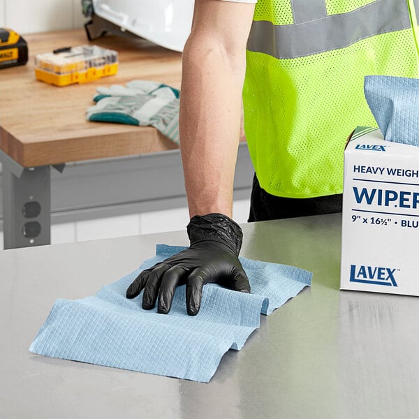 A person in a safety vest and black gloves using a Lavex blue heavy weight industrial wiper to clean a table.