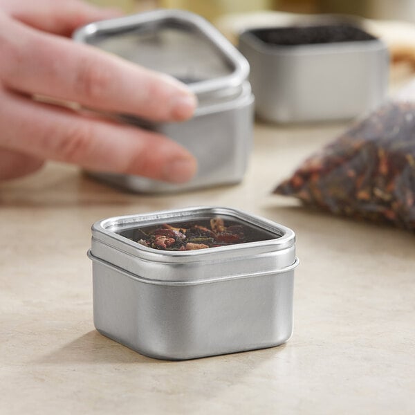 A hand holding a silver square tin with a clear window lid containing herbs.