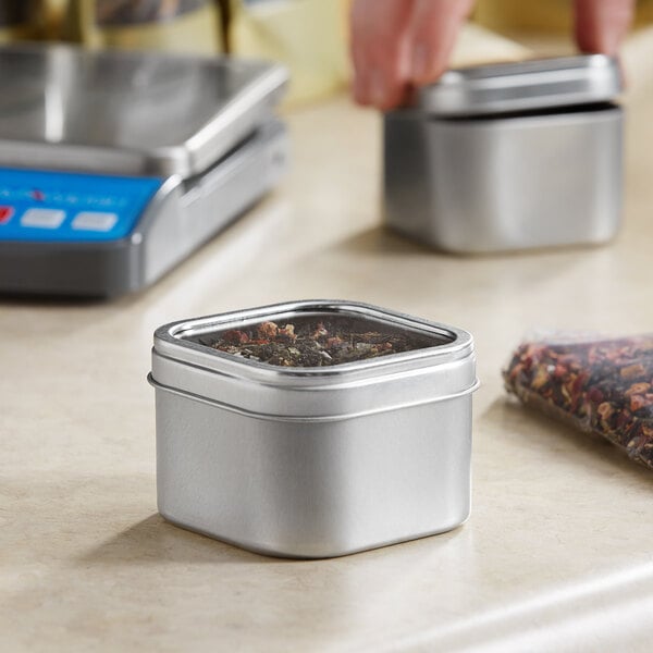 A person using a measuring device to fill a silver square tin with clear window lid with herbs.