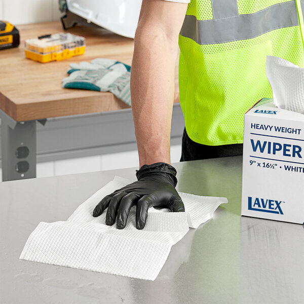 A person wearing a safety vest and black gloves using a Lavex white industrial wiper box to clean a table.