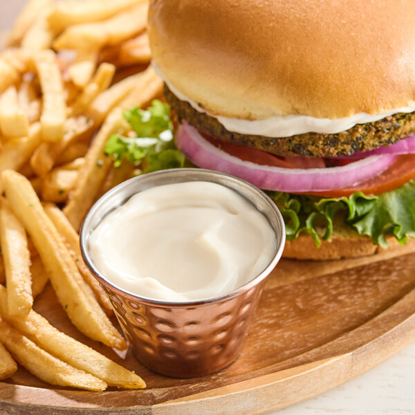 A small metal cup filled with creamy vegan mayonnaise is placed next to French fries and a veggie burger on a wooden plate.