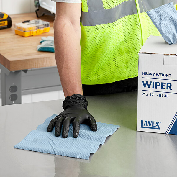 A man in a safety vest and black gloves pulls a blue industrial wiper from a box on a table.