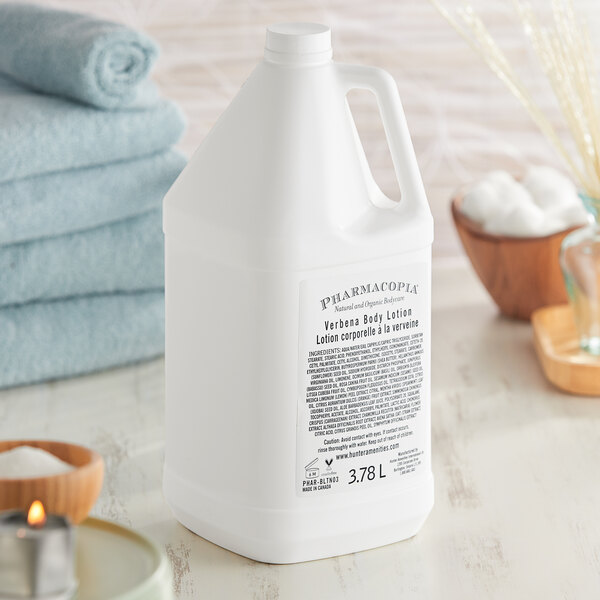 A white jug of Pharmacopia Verbena body lotion on a towel next to a lit candle.