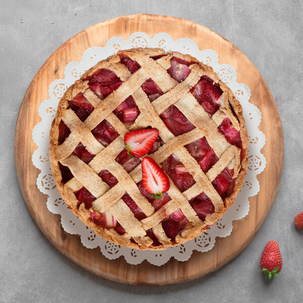 A round pie with a lattice crust sits on a decorative white lace doily atop a wooden board, with strawberries nearby.