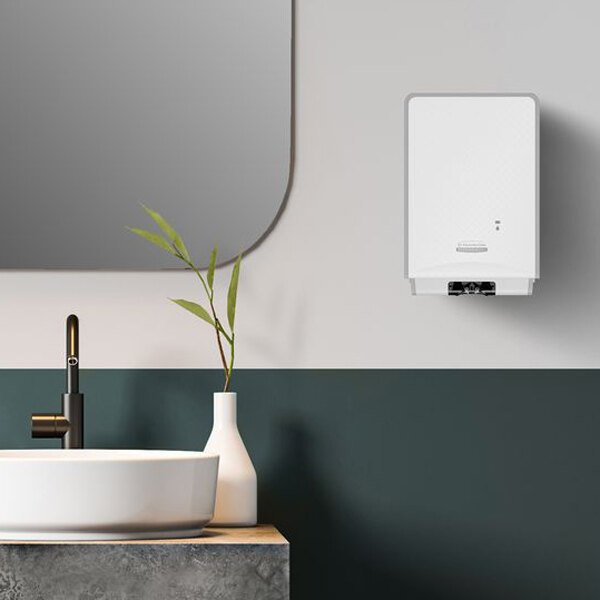 A modern bathroom scene featuring a round white sink, a black faucet, a white vase with green leaves, a wall-mounted rectangular mirror, and a white wall-mounted dispenser.