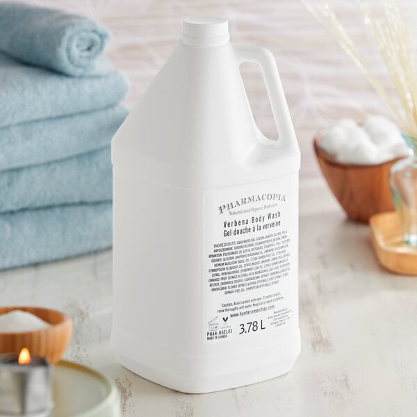 A white plastic bottle of Pharmacopia Verbena body wash on a table next to blue towels and a candle.