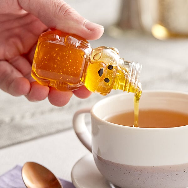 A hand pouring honey from a small yellow bear bottle into a cup of tea.