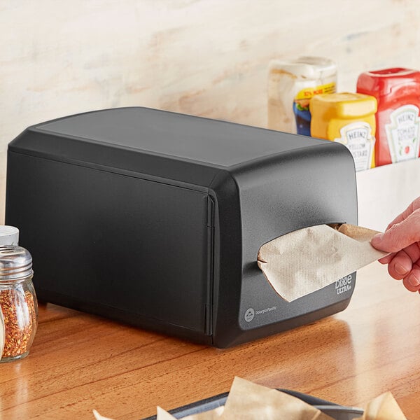 A person putting a black rectangular Dixie Ultra napkin into a black countertop napkin dispenser.