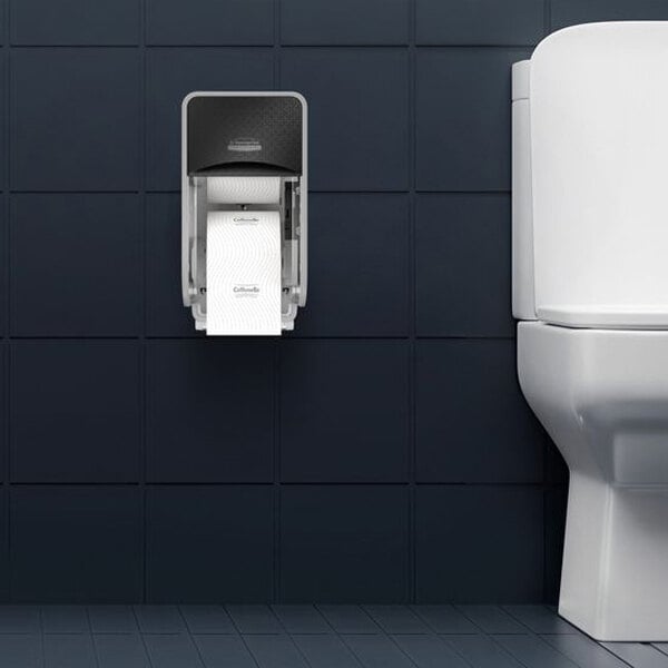 A wall-mounted vertical toilet paper dispenser is installed next to a white toilet against a dark tiled wall.