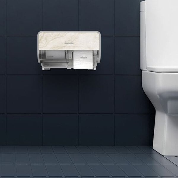 A wall-mounted toilet paper dispenser with a marble-like cover is installed next to a white toilet against dark tiled walls and floor.
