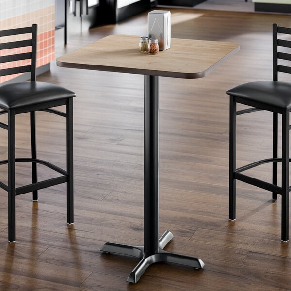 A square bar height table with a light wood laminate top and a black cast iron base.