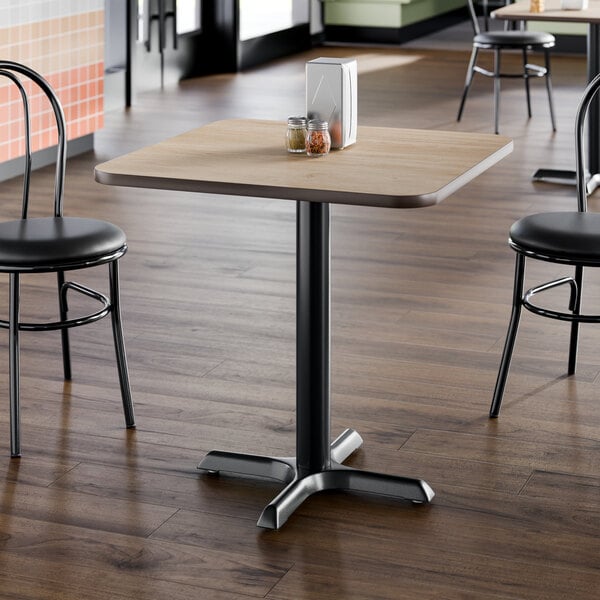 A square laminated restaurant table with a light wood finish and a black cast iron pedestal base.