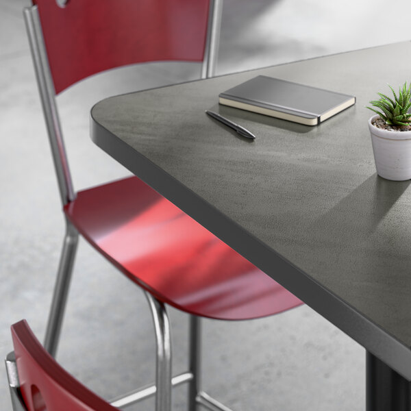 A square bar-height table with a gray laminated top, paired with red metal chairs and a small potted plant, notebook, and pen on the table.