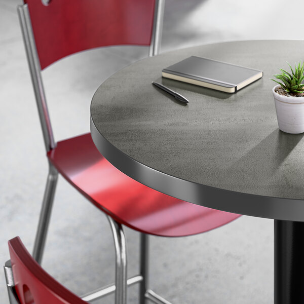 A round gray laminated table with a black edge, accompanied by red chairs, a small potted plant, a notebook, and a pen on top.