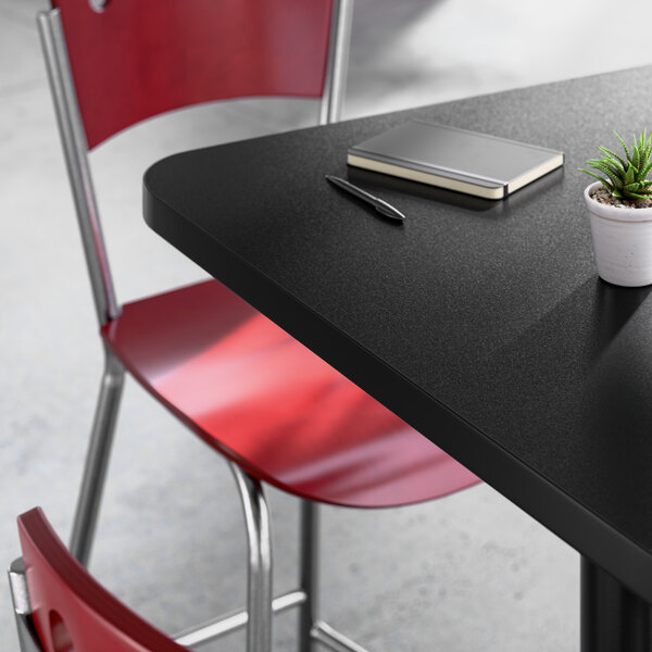 A square black laminated table with a cast iron base, accompanied by a red metal chair, a notebook, a pen, and a small potted plant on top.