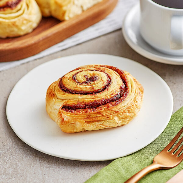 A plate with a Orange Bakery cinnamon roll on it next to a cup of coffee.